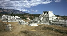 Un grup de piràmides esglaonades blanques, la més alta coronada amb un santuari amb tres entrades. Al fons, una carena de turons.