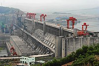 Aerial view of a huge concrete structure located between the mountains, with buildings, power lines and construction and lifting cranes