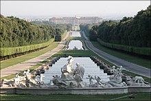 Frontale Farbfotografie einer rechteckigen Wasserfläche, die bis zum Hintergrund führt und an einem Schloss endet. Das Wasser wird durch zwei kleine Rasenflächen durchbrochen. An beiden Seiten sind Wege, Heckenreihen und Wälder. Im Vordergrund sind mehrere Skulpturen, die eine Szene darstellen.