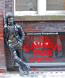 A statue depicting a young Lennon outside a brick building. Next to the statue are three windows, with two side-by-side above the lower, which bears signage advertising the Cavern pub.