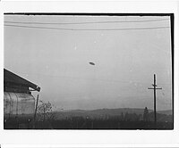 Photographie en noir et blanc d'un objet en forme de disque sur fond de ciel. Bâtiments à proximité.