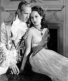 Photographie en noir et blanc de Leslie Howard et Merle Oberon en costume de tournage. Ils sont assis côte à côte, et les regard sont croisés: Oberon, à gauche, regarde au loin vers la droite et Howard, à droite, regarde vers la gauche.