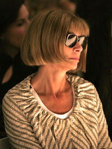 Anna Wintour con gafas de sol y un top de rayas grises y blancas sobre un fondo oscuro mirando a la derecha