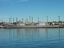 A small warship tied up alongside at a wharf. Her communications and radar masts have been crudely downsized, she carries no weapons, and several large squares have been cut into the ship's hull.