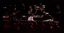 Singer Bruce Springsteen and several other musicians onstage with a placard in front of Springsteen on which is written "I'm Goin' Down"