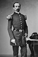 Black and white photo shows a slim man with a moustache and Van Dyke beard. He is standing and wearing a dark military uniform with large epaulettes.