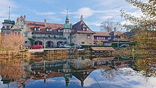 Das Bild zeigt den Hof Riedern, eine Villa die im Stil eines Gutshofs des 19. Jahrhunderts gebaut wurde und die Spiegelung des Gebäudes in einem davor liegenden Weier.