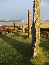 Four large standing stones sit in a field of grass and heather. They are illuminated by reddish sunlight and they cast long shadows to the left. A lake and low hills lie beyond.