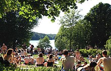 Farbfotografie mit Blick auf eine Wiese mit vielen Badegästen, von denen die meisten in Rückenansicht sind. Im Hintergrund sind der See und viele Bäume.