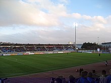 Vue du terrain de Laugardalsvöllur depuis les tribunes du stade. Sous un ciel bleu voilé par des nuages, le terrain est bordé par une piste d'athlétisme. Les tribunes opposées du stade sont presque pleines.