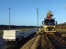 A road and a railway track next to each other, the latter at a higher grade than the former. Overhead lines supported by green metal posts on the left are suspended over the track. A telephone wire supported by a wooden pole on the left runs across the image, above the overhead lines. On the side of the road next to the track is a white pickup truck with a large box on the back; red and yellow striped posts are along the road to its left. On the track is a road-rail vehicle, front facing the viewer, with its lights on. Two men wearing neon green safety clothing with reflectorized stripes are in a fully retracted cherry picker on top