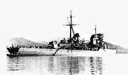 Water-level rear quarter view of a large grey warship at anchor. One gun turret, the main mast and both funnels are prominent.