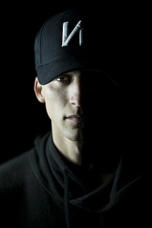 Portrait photo of a man wearing a brimmed cap sporting the "NF" logo and a black hoodie, his entire left side obscured in dark shadow