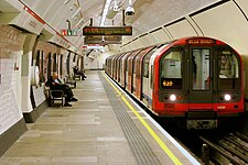 A deep level train stops to the right of a platform as some people (چپ) wait to board it.