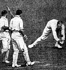 A cricket fielder picks up a ball, watched by two other players