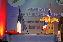 Photographie d'une estrade de part et d'autre de laquelle peuvent être vus le drapeau français et le drapeau du Royaume-Uni et en arrière plan le nom des deux communes jumelées des deux pays.