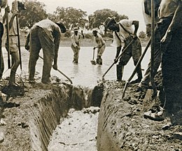 Een zwart-witfoto van mensen die een sloot met stilstaand water dempen