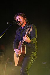 A man semi-inclined with semi-long hair, wearing black jacket and pants, holding a guitar in both hands, apparently singing into a microphone with stand.