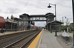 A train station with two tracks and two platforms on each side, connected by an overhead glass bridge