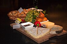 la photographie couleur représente une nature morte. Une planche à découper en bois sert de support à trois fromages. Des olives dans un ramequin, des poires, un poivron et une grappe de raisin blanc complètent la scène. En arrière-plan, une corbeille en osier contient des petits pains ronds