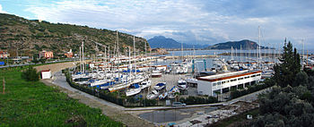 Dozens of sailboats crowd a marina under a cloudy sky along a mountain-lined coast.