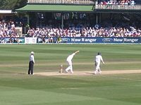 A bowler, midway through delivery. An umpire with black trousers and a light shirt watches from the left while the batsman not receiving the delivery is already walking down the pitch. In the background is a two-tier stand, full of spectators.