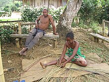 Une jeune malgache assise sur le sol, tresse des roseaux, pendant que son père est assis sur un banc à l'arrière.