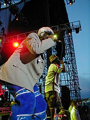 Two members of a musical group, on-stage. The nearest is wearing blue trousers and a gray jacket, while the farthest is wearing shorts, a short-sleeved shirt and a hat.