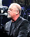 A man with a goatee and his eyes closed behind a microphone on a piano, wearing a black shirt and striped jacket.