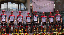 8 male cyclers dressed in red, black, and white uniforms with baseball caps, standing on a stage mounting their bikes. A man fourth from the right is waving his hand towards a crowd not depicted, with a line of flower bouquets in yellow, orange, and red lining the bottom of the picture.