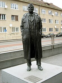 A dark bronze statue of a man looking straight forward with his hands in his coat pockets.