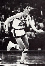 A man, wearing a black suit and a tie, is making a gesture by raising his hands while standing on the side of a basketball court.