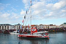 Un voilier rouge et blanc, voiles baissées, descend le chenal des Sables d'Olonne. Une foule se presse sur le quai.