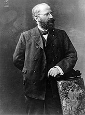 Eduard Bernstein standing next to a chair and looking rightward. He is resting his hand on the chair.