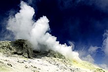 Fumarolas blancas escapando de grietas en la cima de una montaña con depósitos de azufre de color amarillo pálido a amarillo a su alrededor, bajo un cielo azul intenso.