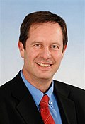 Man with dark hair looking straight ahead, head and shoulders shot, dark jacket, blue shirt, red tie.