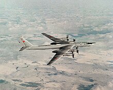 Top perpendicular starboard view of four-engine propeller-driven aircraft in-flight. The pale gray/white aircraft is flying above almost white terrain.