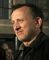 Headshot of a middle-aged man looking to his right. The man has short hair and light stubble. He appears to be smiling slightly, and is wearing a dark coat.