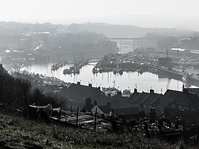 A black and white image looking upriver from Whitby Abbey. It shows the wide estuary harbour with many pleasure boats in the marina