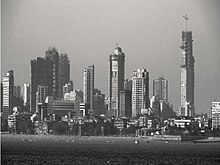 Skyscrapers under construction dotted by maller buildings and trees. A beach is in front of them