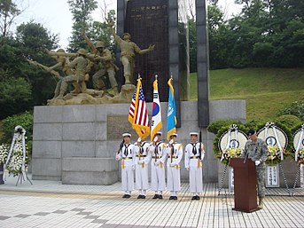 A line of sailors in white dress naval uniforms in front of a large monument while another soldier in green stands at a podium beside