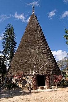 Maison conique avec un toit de chaume, au sommet une flèche en bois sculpté.