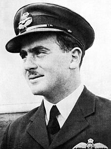 Informal head-and-shoulders portrait of a dark-haired, moustachioed man in dark military jacket with pilot's wings on the left breast pocket, and peaked cap