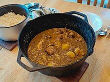 A cast-iron pot of chicken and potato xacuti, a warm yellow-brown curry, sits on a table. There is a small pot of rice visible in the top right of the frame.