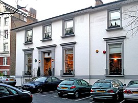 A two storey white building with grey window frames, several cars are parked in the foreground.