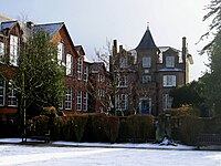 A colour photograph of an unusual Victorian house with a small spire on the top. To the left is a set of old-fashioned schoolrooms with large sash windows. In front of the house is a small lawn, covered in snow.