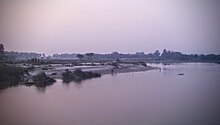 Figures stand on the sandy far bank of a slow-flowing river.