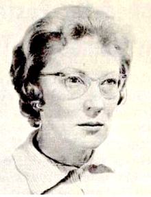 A middle-aged white woman with short wavy hair, wearing glasses and a collared top buttoned to the throat