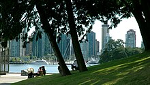 sebuah bukit berumput hijau dititik dengan cerunan pokok bawah ke suatu kawasan diterap dengan bangku-bangku. Beyond lies water, docks, and a yacht, and skyscrapers.