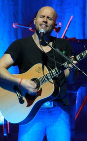 A bald Peruvian man, wearing a black shirt and blue jeans, holding an acoustic guitar with a microphone in front of him.